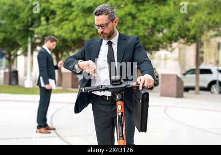 Homme d'affaires attrayant conduisant un scooter de coup de pied au parc de New York. Homme d'affaires dans un costume chevauchant un scooter électrique sur une réunion d'affaires. Âge moyen Banque D'Images