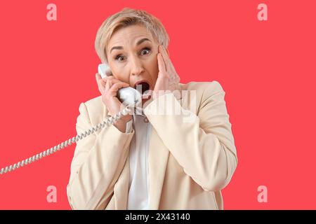Femme mature choquée parlant par téléphone sur fond rouge Banque D'Images