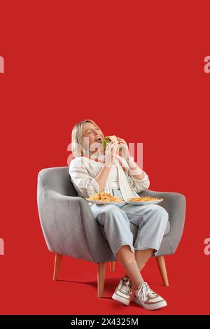 Belle femme avec de la nourriture malsaine assise dans un fauteuil sur fond rouge. Concept de suralimentation Banque D'Images