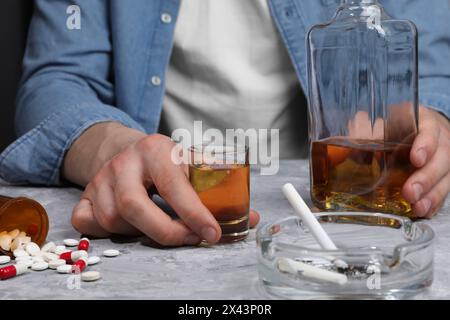 Alcoolisme et toxicomanie. Homme avec whisky, pilules et cigarettes fumantes à la table texturée grise, gros plan Banque D'Images