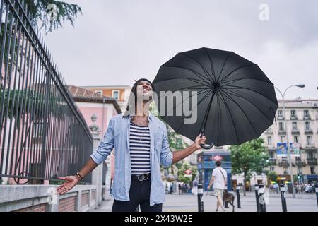 Jeune homme attrayant sous la pluie, tenant son parapluie, levant les yeux vers le ciel et se sentant joyeux et heureux Banque D'Images