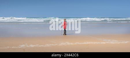 Une image panoramique d'une femme mature portant un manteau rouge vif debout seule sur le rivage à la plage de Mawgan Porth en Cornouailles au Royaume-Uni. Banque D'Images