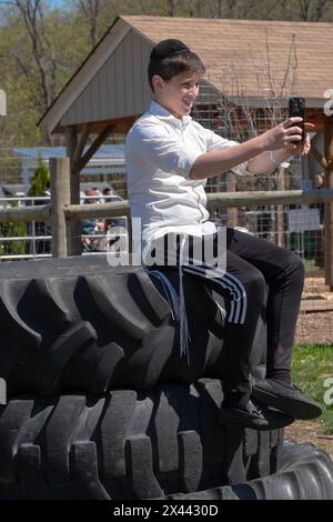 Un adolescent juif orthodoxe prend un selfie assis sur une pile de pneus surdimensionnés. Dans une ferme du comté de Rockland et pendant la Pâque 2024. Banque D'Images