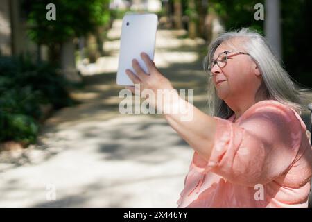 Vieille dame positive prenant selfie dans le parc. Femme aux cheveux gris senior dans le gadget décontracté tenant avec la main et regardant la caméra du téléphone. Concept d'auto-prise de vue Banque D'Images