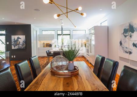 Table à manger en bois de rose avec chaises à haut dossier en cuir brun foncé dans la salle à manger avec parquet en bois d'acacia et vue sur la cuisine avec blanc brillant Banque D'Images