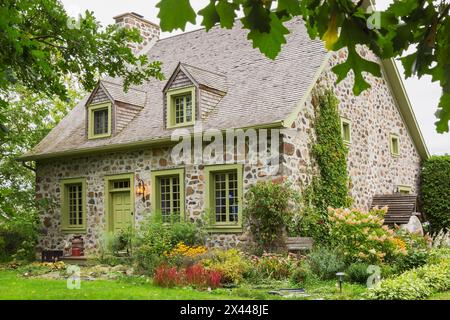 Ancienne façade de maison en pierre de champ Canadiana vers 1735 avec garniture vert citron et toiture en bardeaux de bois de cèdre et cour avant paysagée à la fin de l'été, Québec Banque D'Images