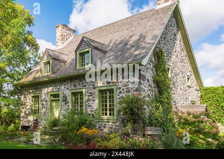 Ancienne façade de maison en pierre de champ Canadiana vers 1735 avec garniture vert citron et toiture en bardeaux de bois de cèdre et cour avant paysagée à la fin de l'été, Québec Banque D'Images