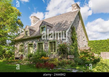 Ancienne façade de maison en pierre de champ Canadiana vers 1735 avec garniture vert citron et toiture en bardeaux de bois de cèdre et cour avant paysagée à la fin de l'été, Québec Banque D'Images