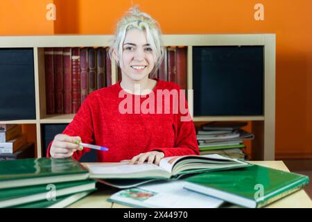 Une jeune femme joyeuse aux cheveux blonds, dans un pull rouge, souriant à la caméra tout en tenant un stylo, assise à un bureau dans une bibliothèque entourée de livre Banque D'Images