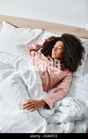 Une femme afro-américaine bouclée en pyjama reposant paisiblement sur un lit blanc dans une chambre ensoleillée pendant l'heure du matin. Banque D'Images