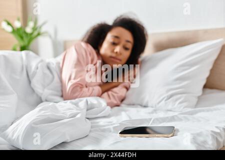 Une femme afro-américaine en pyjama est couchée dans son lit, absorbée dans son téléphone portable dans la douce lumière du matin. Banque D'Images