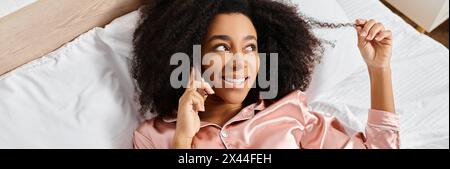 Femme afro-américaine bouclée en pyjama couché dans le lit, ayant une conversation sur son téléphone portable le matin. Banque D'Images