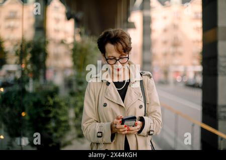 Femme âgée retraitée utilisant un téléphone intelligent tout en marchant sur le trottoir Banque D'Images