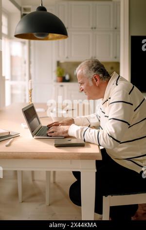 Vue latérale de l'homme senior retraité utilisant un ordinateur portable à la table à manger dans la cuisine Banque D'Images