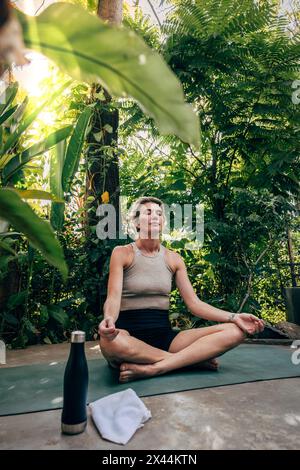 Femme pratiquant la position Lotus tout en étant assis sur le tapis de yoga près des plantes à la station de bien-être Banque D'Images