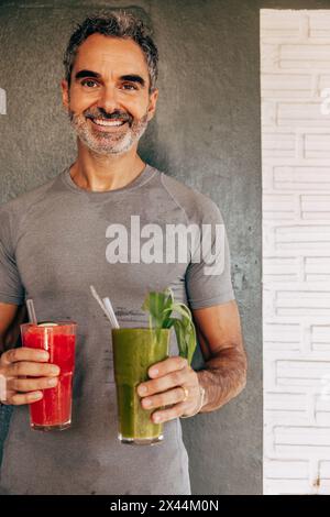 Portrait d'un homme mature souriant tenant des verres à smoothie tout en se tenant debout contre le mur à la station de bien-être Banque D'Images