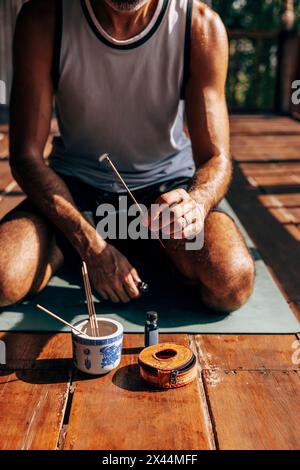 Section basse de l'homme tenant le bâton d'encens brûlant tout en étant assis sur le tapis de yoga à la station de bien-être Banque D'Images