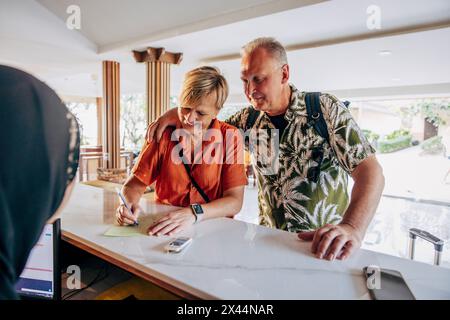 Femme senior souriante remplissant le formulaire avec un homme tout en se tenant debout à la réception de l'hôtel Banque D'Images