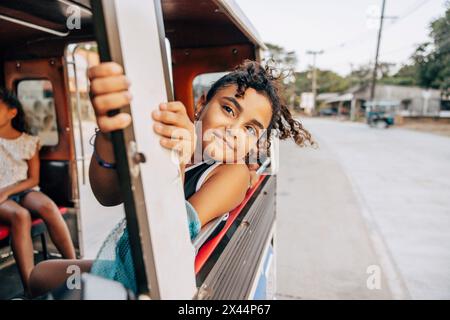 Fille souriante se penchant par la fenêtre pendant le voyage sur la route tout en étant assis dans tuk-tuk en vacances Banque D'Images