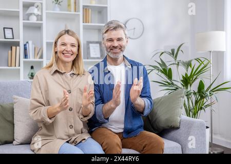 Un joyeux couple d'âge mûr assis sur un canapé, applaudissant les mains avec de larges sourires dans un salon élégamment décoré. Exprimer le bonheur et le contentement. Banque D'Images