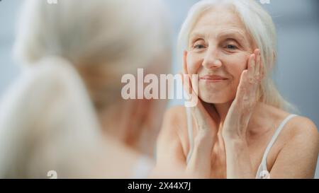 Belle femme senior regarde dans la salle de bain miroir touche son visage, joyeusement sourit, profiter de son apparence et de la routine du matin. Dignité et grâce dans la vieillesse. Bien-être, soins de la peau, produits de beauté Banque D'Images