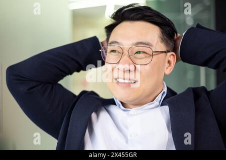 Homme d'affaires asiatique d'âge moyen se relaxant avec ses mains derrière la tête, souriant dans un environnement de bureau lumineux. Il respire le contentement et la confiance. Banque D'Images