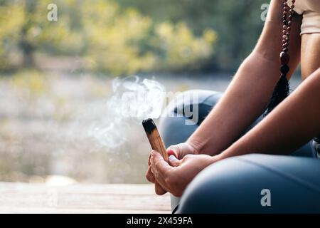 Gros plan d'une femme méconnaissable en vêtements de sport méditant et faisant du yoga dans la nature. Elle brûle un palo santo en bois. Banque D'Images