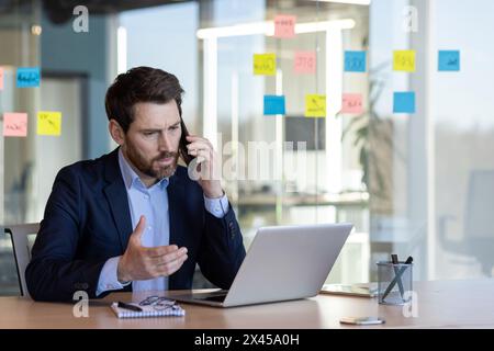 Un homme d'affaires professionnel en costume s'engage activement dans une conversation téléphonique tout en regardant son ordinateur portable dans un environnement de bureau moderne. Banque D'Images