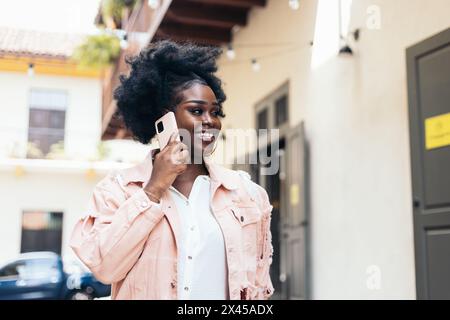 Femme noire élégante avec cheveux afro marchant dans la rue de la ville tout en parlant sur le téléphone portable et en riant. Elle porte des vêtements décontractés et de grandes boucles d'oreilles Banque D'Images