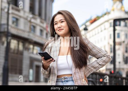 Jeune femme chinoise adulte dans une veste à carreaux décontractée à l'aide d'un smartphone dans les rues de la ville européenne Banque D'Images