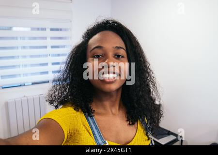 Smiling Selfie : jeune femme noire avec de longs cheveux bouclés en chemise jaune faisant des selfies à la maison Banque D'Images