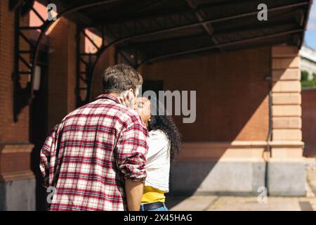 Couple de jeunes adultes multiraciaux embrassant et embrassant après une réunion à nouveau à la gare. Elle porte une valise bleue de son voyage et il attend Banque D'Images