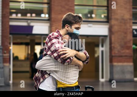Couple de jeunes adultes multiraciaux embrassant et embrassant après une réunion à nouveau à la gare. Elle porte une valise bleue de son voyage et il attend Banque D'Images