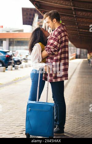 Couple de jeunes adultes multiraciaux embrassant et embrassant après une réunion à nouveau à la gare. Elle porte une valise bleue de son voyage et il attend Banque D'Images