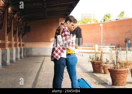 Couple de jeunes adultes multiraciaux embrassant et embrassant après une réunion à nouveau à la gare. Elle porte une valise bleue de son voyage et il attend Banque D'Images