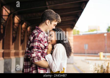 Couple de jeunes adultes multiraciaux embrassant et embrassant après une réunion à nouveau à la gare. Elle porte une valise bleue de son voyage et il attend Banque D'Images