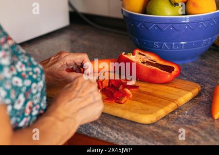 Gros plan d'une femme âgée avec des verres préparant un délicieux repas à la maison. Elle coupe des poivrons rouges dans la cuisine Banque D'Images