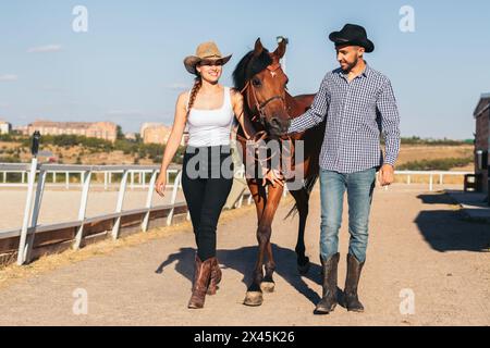 Jeune homme et femme avec des chapeaux et des bottes de style cow-boy marchant avec leur cheval dans un centre équestre à la campagne. Banque D'Images