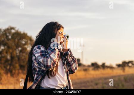 Jeune femme en chemise à carreaux et vêtements décontractés prenant des photos avec un appareil photo analogique dans le champ au coucher du soleil. L'image véhicule le concept de rétro a Banque D'Images