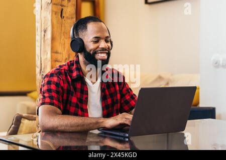 Homme noir souriant travaillant à distance depuis la maison avec un ordinateur portable et des écouteurs. Concept de télétravail et de bureau à domicile. Banque D'Images