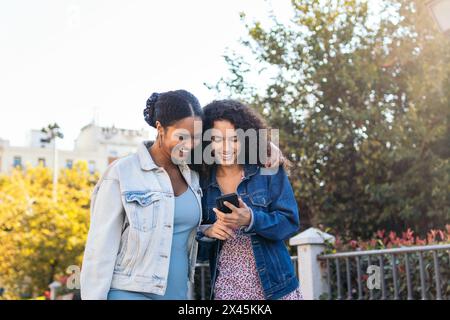 Deux amis latino-américains marchant dans la rue, s'amusant et riant en utilisant un smartphone. Banque D'Images