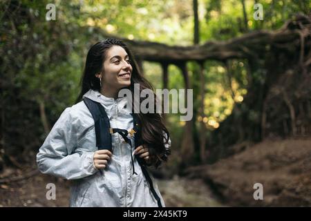 Jeune femme avec imperméable et sac à dos marchant à travers une jungle avec de grands arbres. Banque D'Images