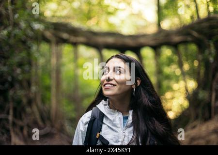 Jeune femme avec imperméable et sac à dos marchant à travers une jungle avec de grands arbres. Banque D'Images