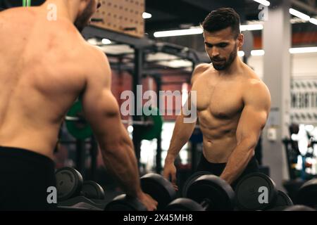 homme fort debout devant le miroir prenant des haltères dans une salle de gym Banque D'Images