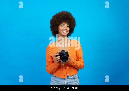 Portrait d'une photographe avec des cheveux afro avec appareil photo dans ses mains sur fond bleu Banque D'Images