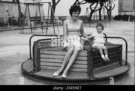 Années 1960, historique, été et une mère dans une robe sans manches à motifs floraux assise avec son fils bébé sur le siège d'un rond-point traditionnel en bois, encadré de métal dans une aire de jeux en bord de mer, Malte. Banque D'Images