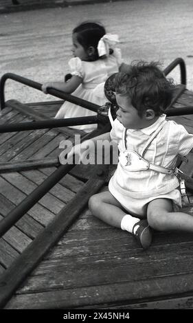 années 1960, historique, un enfant en bas âge, portant un harnais de sécurité en cuir, assis ayant une promenade sur la plate-forme en bois surélevée d'un rond-point traditionnel de terrain de jeu en métal. Banque D'Images