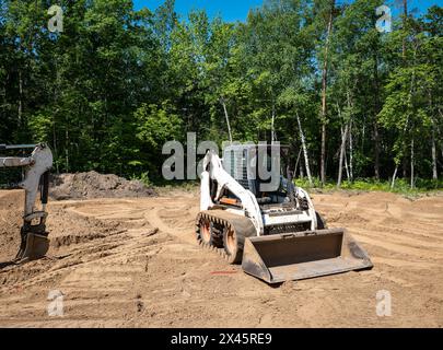 Chargeur frontal compact blanc, avec chenilles de mini-chargeuse, repose sur la terre dans un terrain vide bordé d'arbres, en préparation de la construction d'une nouvelle maison. Le Banque D'Images