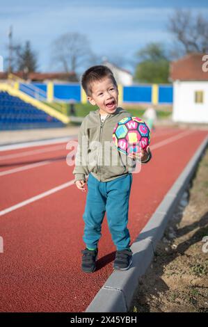 Un jeune garçon joyeux saisit un ballon de football dynamique sur la piste, signalant l'intérêt de l'enfant de 3 ans pour le football dès son plus jeune âge Banque D'Images