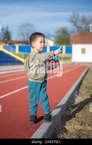 Un jeune garçon joyeux saisit un ballon de football dynamique sur la piste, signalant l'intérêt de l'enfant de 3 ans pour le football dès son plus jeune âge Banque D'Images